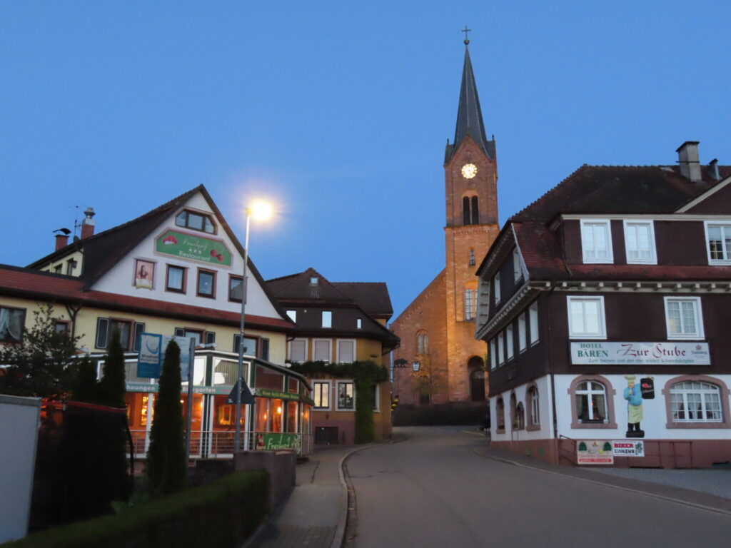 Abendstimmung in einem idyllischen Dorf im Schwarzwald: Im Vordergrund traditionelle Gasthäuser wie das Hotel „Zur Stube Bären“ und ein Restaurant mit der Aufschrift „Frühauf“, dahinter ragt der beleuchtete Kirchturm der Dorfkirche hervor. Eine ruhige Straße führt durch die Szene, umgeben von Fachwerkhäusern und Laternenlicht.